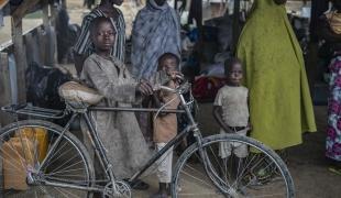Un jeune garçon et sa bicyclette dans le camp de transit de la ville de Gwoza, où les personnes déplacées attendent de recevoir une tente familiale. 2018. Nigeria. 