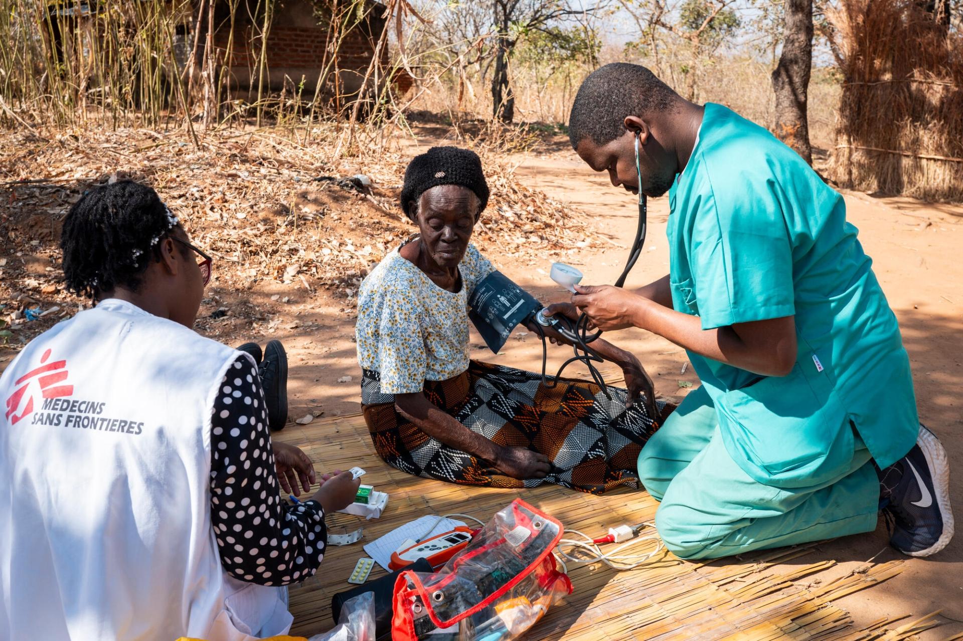 Des membres de l'équipe MSF prennent la tension artérielle de Grace Nyambi, 72 ans. Prise en charge depuis avril 2025, elle souffre de douleurs abdominales ; l'équipe lui apporte de la morphine et des patchs antidouleur.