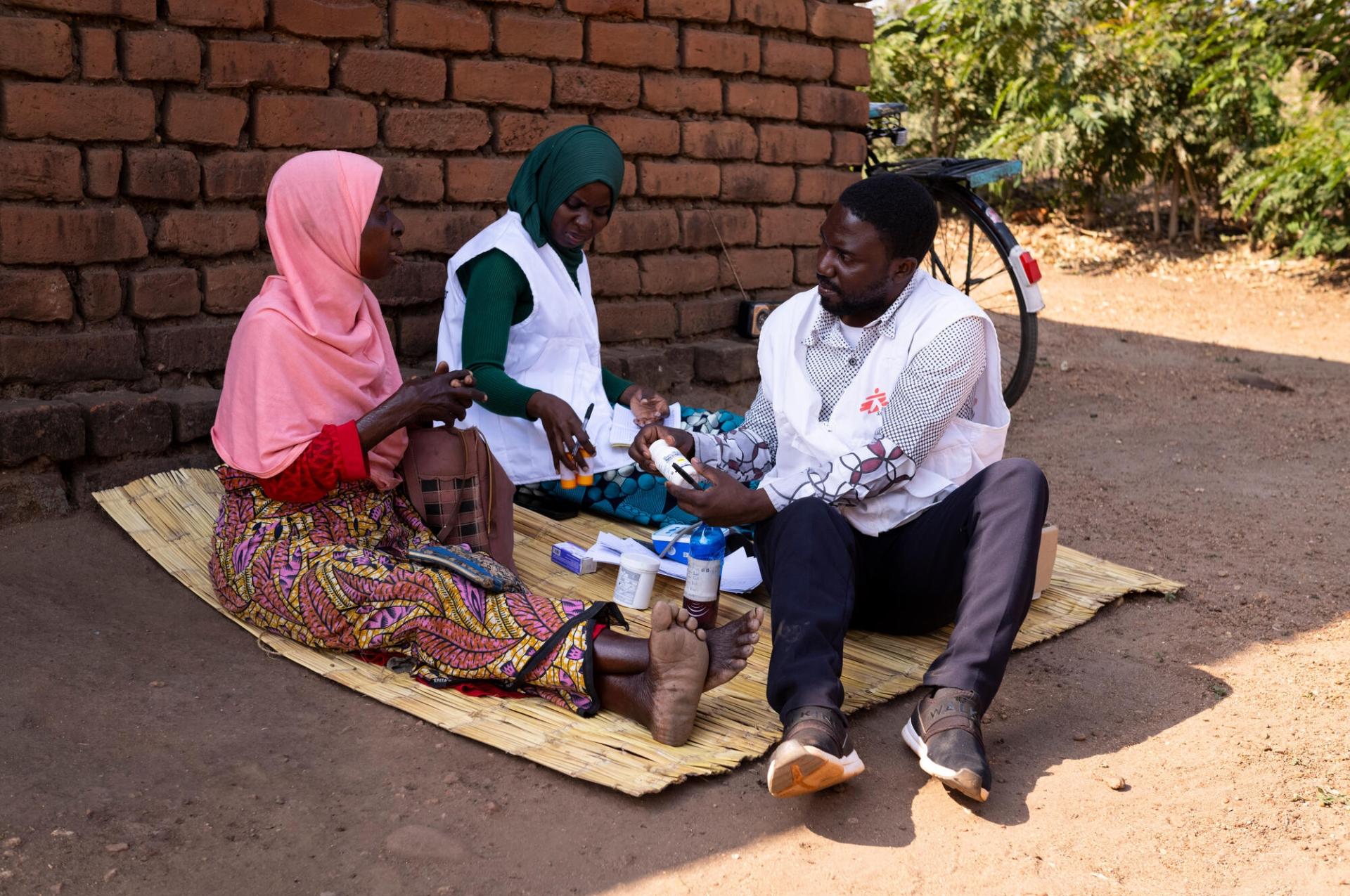 Une patiente dans son village avec deux membres de l'équipe de MSF qui lui apportent une aide médicale face à son cancer.